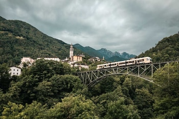 Un tren blanco y amarillo cruza un puente de acero sobre un río en un valle montañoso cubierto de densa vegetación verde, con nubes bajas en las cimas
