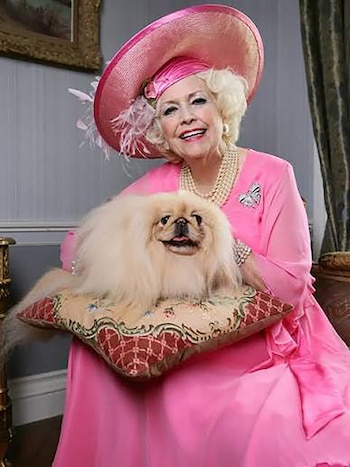 Barbara Cartland con sombrero y vestido rosas sonríe junto a un perro pequinés recostado en un cojín