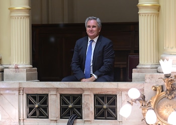 Hombre de mediana edad con traje oscuro y corbata azul sentado en un balcón interior de mármol, flanqueado por columnas estriadas y una lámpara ornamental
