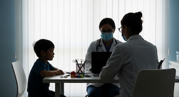 Un niño, dos profesionales médicos con batas y mascarillas, una computadora portátil, tijeras, lápices, juguetes y pastillas sobre un escritorio.