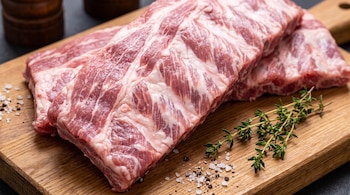 Dos costillas de cerdo crudas en una tabla de cortar de madera, con sal gruesa, pimienta negra molida y ramitas de tomillo fresco.