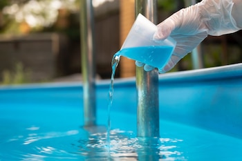 Una mano con guante transparente vierte líquido azul desde un vaso medidor transparente hacia el agua azul de una piscina, con una barandilla metálica visible
