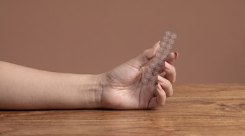 Una mano de mujer sostiene una tira transparente con parches circulares adhesivos. La mano está sobre una superficie de madera. Hay un fondo liso de color marrón.