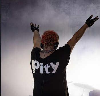 Vista trasera de Pity Álvarez en un escenario, con los brazos alzados, cabello rizado naranja, camiseta negra con 'Pity' en blanco, auriculares y guantes negros