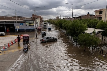 Una calle inundada con agua marrón por la que circulan vehículos, mototaxis y una motocicleta junto a árboles y edificaciones