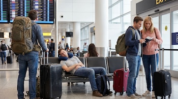 Varios viajeros en una terminal de aeropuerto, incluyendo un hombre con mochila mirando una pantalla y una pareja revisando pasaportes, con equipaje.