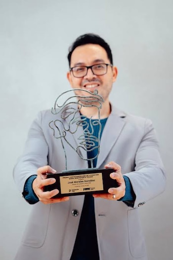 Un hombre con gafas y saco gris sostiene un trofeo metálico con una placa grabada frente a la cámara
