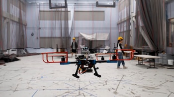 Un dron suspendido en el aire dentro de un laboratorio, con dos personas con cascos amarillos al fondo