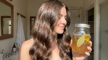 Una mujer sonríe y mira un frasco de vidrio lleno de una infusión amarilla con hojas de laurel, flores de diente de león y lavanda, con su cabello ondulado y brillante.