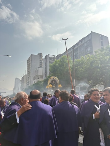 Hombres vestidos con hábitos morados avanzan por una avenida hacia un anda religiosa en una calle con edificios