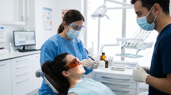 Una paciente con gafas protectoras reclinada en un sillón dental junto a una especialista con pantalla facial y un asistente con mascarilla.