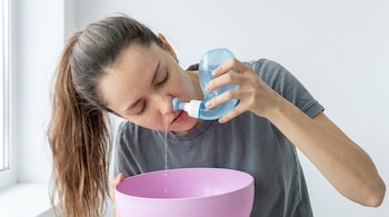 Una mujer inclinada introduce la boquilla de un frasco azul en su nariz mientras el agua cae hacia un recipiente rosado.