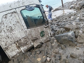 Cabina de camión blanca cubierta de barro y rocas, varada en un terreno lodoso con un hombre caminando hacia el agua en el fondo