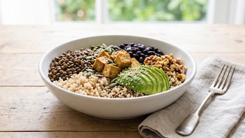 Cuenco blanco con granos cocidos, lentejas, frijoles, tofu, nueces, semillas y aguacate en una mesa junto a una servilleta y un tenedor