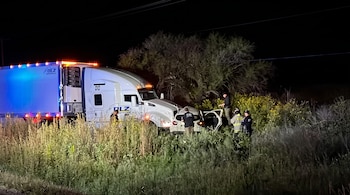 Un camión blanco de transporte y un automóvil compacto detenidos tras un choque en la hierba nocturna junto a personal de socorro.
