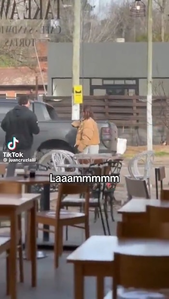 Vista desde un café, un hombre y una mujer caminan junto a una camioneta estacionada. Mesas y sillas de madera en primer plano