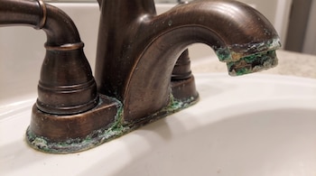 Un grifo metálico oscuro sobre un lavabo blanco con acumulaciones de sarro verde en su base y en el extremo del caño.