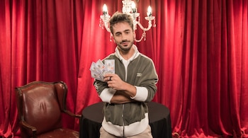 Un hombre con chaqueta verde y blanca sostiene varios billetes de cien dólares. Detrás hay una cortina roja, un candelabro y una mesa.