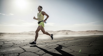 Hombre corre en carretera agrietada con auriculares, botella de agua, camiseta y pantalones cortos. Hay dunas de arena al fondo bajo el sol.