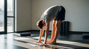 Un hombre con camiseta gris y pantalones cortos azules realiza una flexión de pie hacia adelante sobre una esterilla de yoga oscura en un estudio con luz natural.
