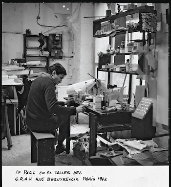 Foto en blanco y negro de Julio Le Parc sentado en su taller, trabajando en una mesa llena de materiales y herramientas, con estanterías atestadas al fondo