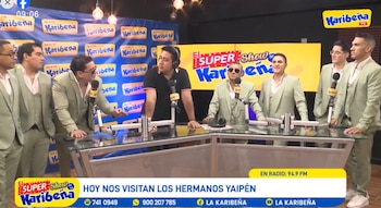 Ocho hombres en un estudio de radio: seis con trajes verdes, dos con ropa informal, junto a micrófonos y una mesa con logos de Karibeña