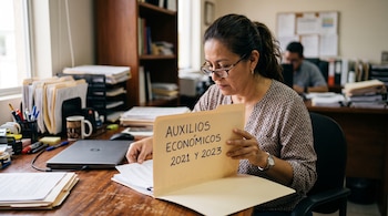 Una mujer con gafas revisa una carpeta amarilla en un escritorio con la inscripción AUXILIOS ECONÓMICOS 2021 Y 2023.