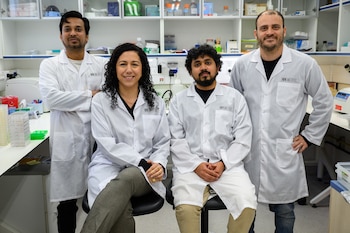 Cuatro científicos de laboratorio médico, tres hombres y una mujer, con batas blancas, posan sonrientes en un laboratorio con estantes y equipos visibles