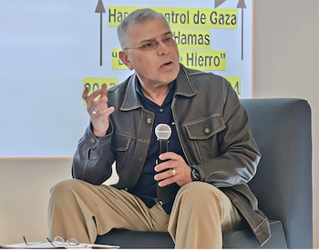 Un hombre de cabello gris y gafas, vestido con chaqueta oscura y pantalones caqui, sentado, hablando a un micrófono; detrás, una pizarra con texto sobre Gaza