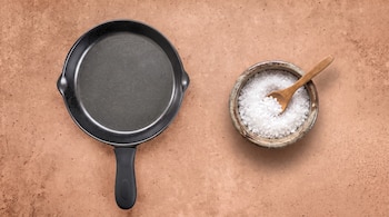 Una sartén negra con mango de goma, un cuenco de cerámica con sal gruesa blanca y una cuchara de madera sobre una mesa marrón.