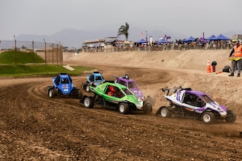 Dos automóviles de carrera transitan por una curva de pista de tierra con agua acumulada y carteles perimetrales al fondo