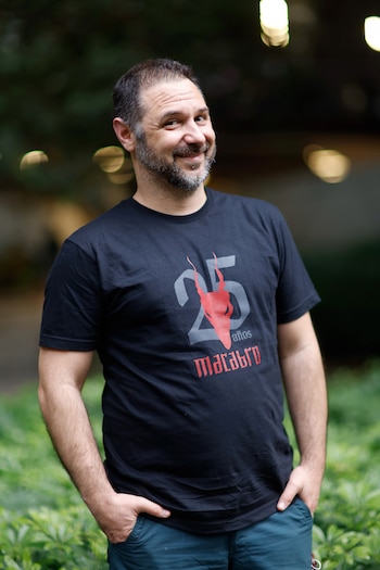 Hombre con barba y camiseta negra con un diseño rojo y gris que levanta ambas manos en un espacio con fondo desenfocado
