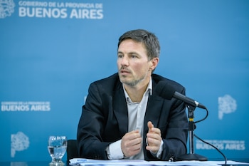 Augusto Costa sentado ante un micrófono y un vaso de agua, con un fondo azul que tiene el logotipo de la Provincia de Buenos Aires