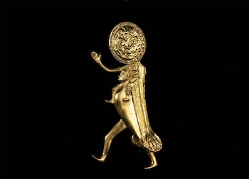 Broche o escultura dorada de una figura humanoide caminando, con un gran rostro solar rodeado de joyas sobre la cabeza y un pequeño busto en el pecho, sobre fondo negro