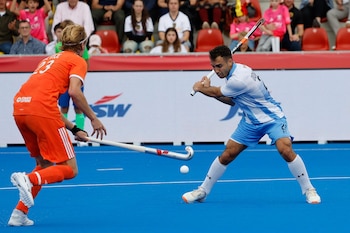 Un jugador de hockey con camiseta celeste y blanca golpea una pelota frente a un rival con uniforme naranja sobre una cancha azul