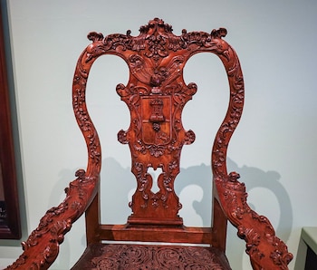 Silla de madera tallada con adornos detallados en los brazos y el respaldo, el cual presenta un relieve de un escudo con una torre