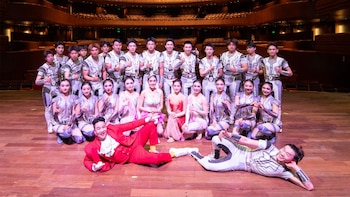 Un grupo de veintiséis acróbatas posa en un escenario de madera; la mayoría viste trajes plateados y blancos, dos artistas yacen en el suelo sonriendo