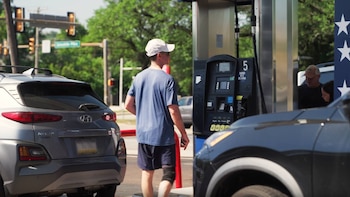Hombre con gorra camina frente a surtidor de combustible. Detrás, SUV Hyundai Kona gris. A la derecha, coche oscuro con dos personas