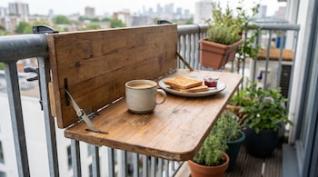 Una mesa abatible de madera, sujeta a la barandilla de un balcón, muestra una taza de café, dos tostadas y mermelada. Se ven plantas en macetas y edificios urbanos al fondo.