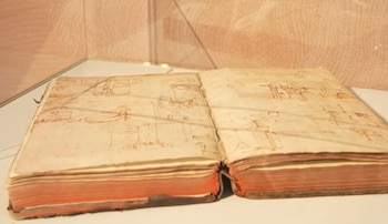 Un códice antiguo abierto en una vitrina. Sus páginas amarillentas muestran bocetos a pluma de mecanismos y escritura. Los bordes del libro son rojizos