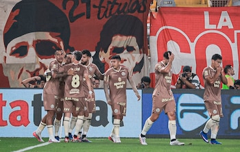 Jugadores de fútbol con uniforme marrón celebran en un campo verde, con pancartas rojas de fondo y fotógrafos