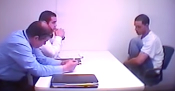 Tres hombres en una sala de interrogatorios, dos sentados a una mesa con documentos y uno sentado enfrente con las rodillas encogidas