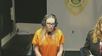 Una mujer con uniforme naranja, anteojos y auriculares con micrófono permanece sentada frente a un fondo con un escudo oficial