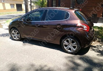 Automóvil Peugeot 208 color marrón oscuro estacionado en una calle de asfalto con casas y vegetación de fondo. Hay sombras en el suelo