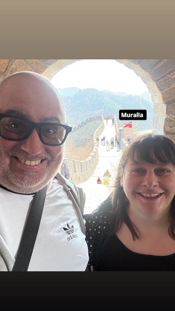 Un hombre y una mujer sonríen en primer plano. Detrás, la Muralla China serpentea por un paisaje montañoso con gente caminando