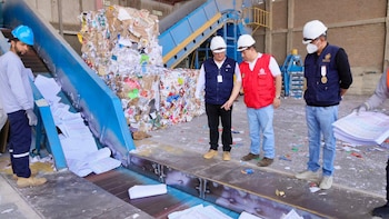 Tres personas con cascos de protección y chalecos observan pliegos de papel en una cinta transportadora dentro de una planta de reciclaje
