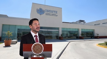 Hombre con traje, corbata y gafas en podio de madera con escudo nacional mexicano. Edificio blanco con logo ISSSTE y cielo azul.