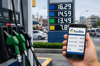Mano sostiene un teléfono móvil con la aplicación Facilito que muestra precios de combustibles frente a surtidores y un cartel digital de precios en una estación de servicio.