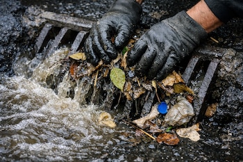 Manos con guantes negros sobre una alcantarilla metálica obstruida por hojas secas, agua turbia, barro y basura como una tapa azul y una bolsa.