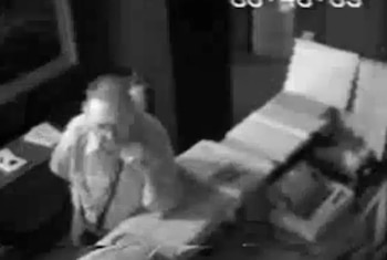 Imagen granulada de CCTV en blanco y negro que muestra a una figura sentada o encorvada en un interior oscuro, con una marca de tiempo digital en la esquina superior derecha